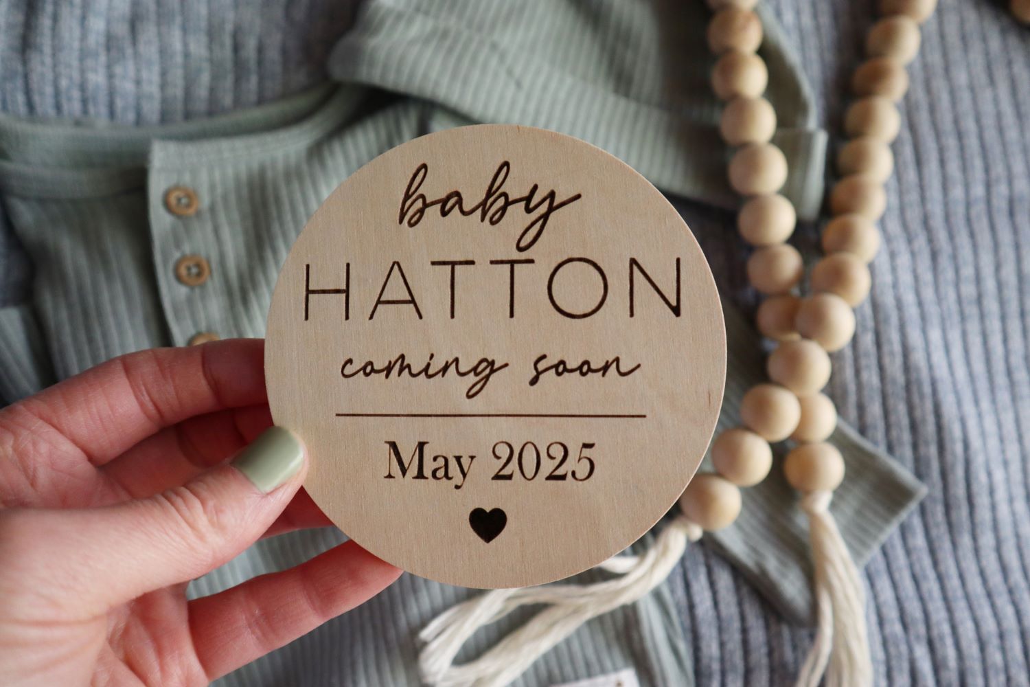 Custom engraved wooden sign reading 'Baby Coming Soon' with last name and due date, displayed against a backdrop of an olive green baby outfit, a baby blue blanket, and wood beads, creating an inviting nursery scene.