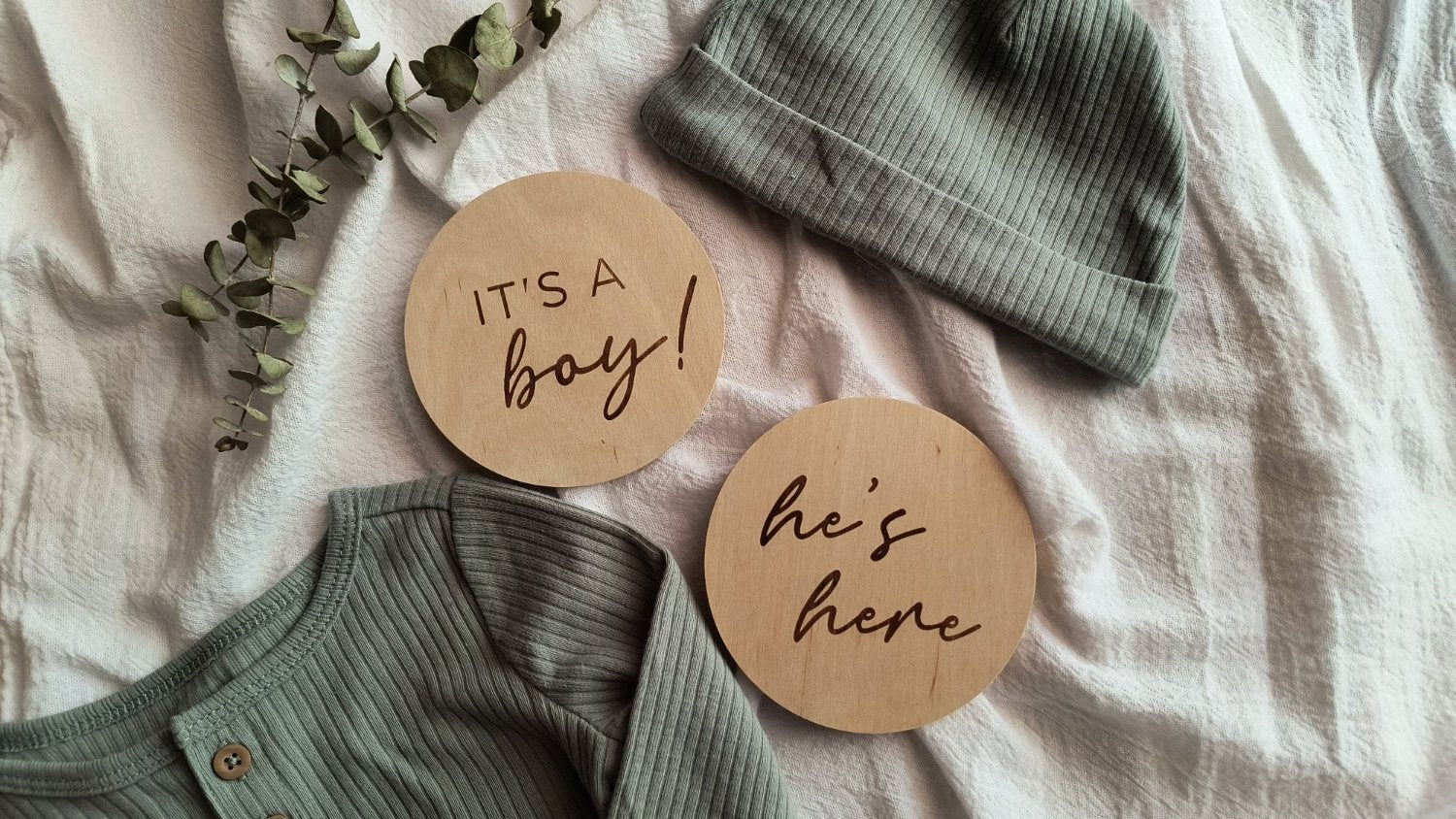 Two engraved wood signs that say 'It's a boy!' and 'he's here' laying on a white blanket with a sage green baby boy outfit and dried eucalyptus greenery to the upper left corner. 