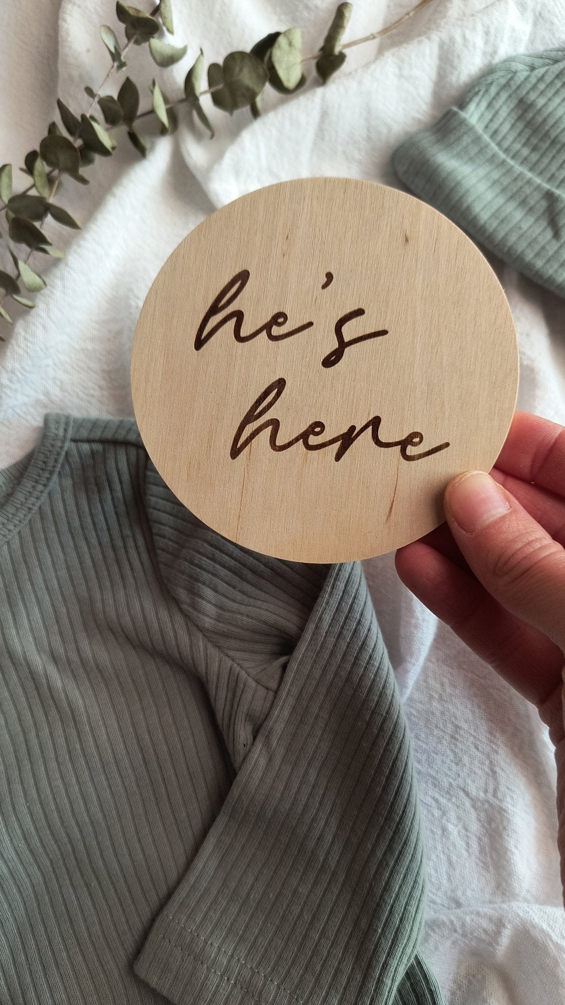 Hand holding engraved 'he's here' gender announcement sign. The background contains a sage green baby boy outfit and eucalyptus leaves. 