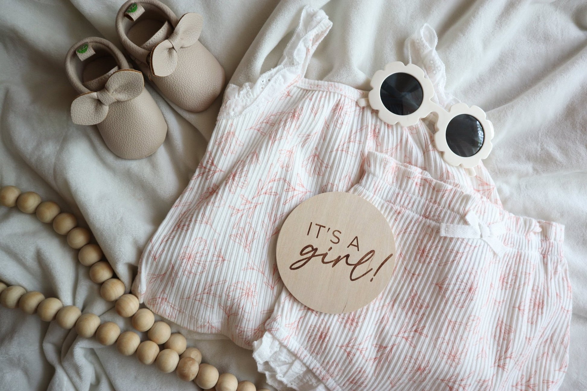 Flatlay of engraved 'It's a Girl!' sign with baby girl outfit, shoes, sunglasses, and wood beads on a white blanket. Perfect for baby announcements and keepsakes.