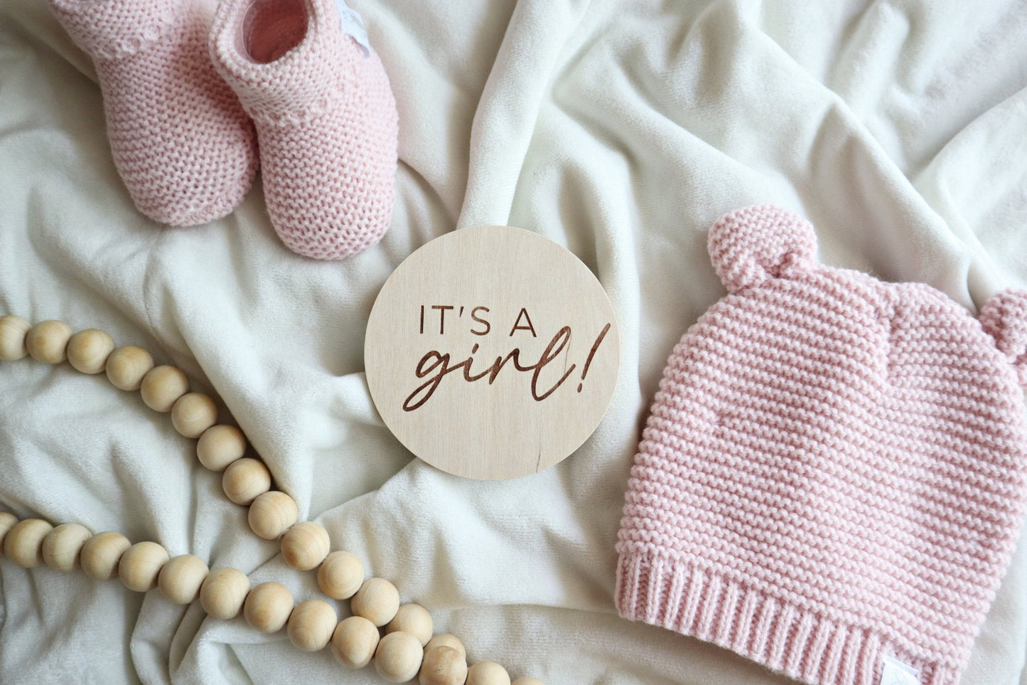 Flatlay of engraved 'It's a Girl!' sign with pink knit baby hat, booties, and wood beads on a white blanket. Perfect for baby announcements and keepsakes.