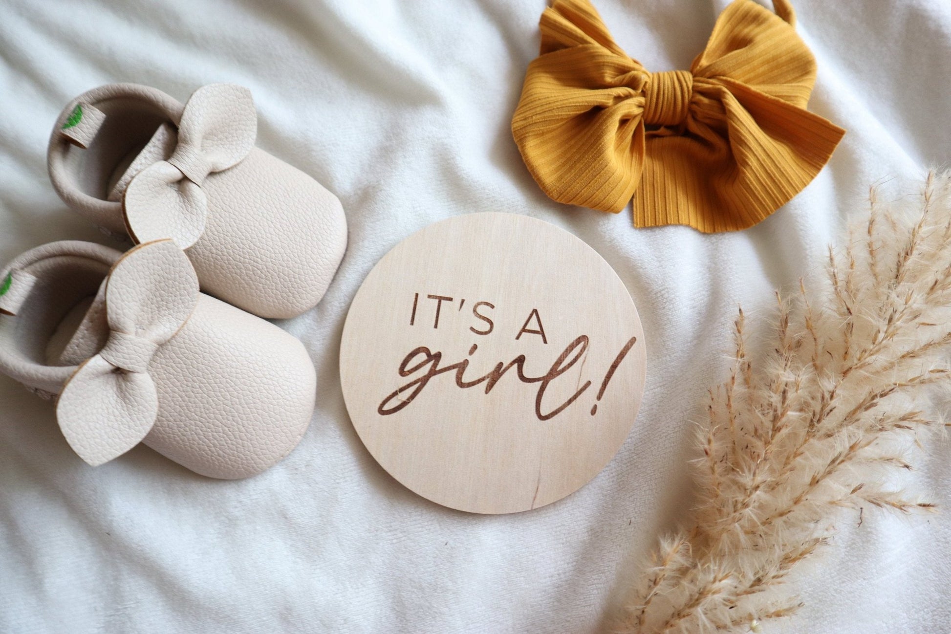 Flatlay of engraved wooden 'It's a Girl!' sign with baby shoes and a bow. Perfect for baby announcement photos and keepsakes.