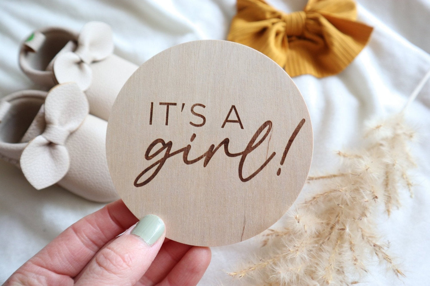 Hand holding an engraved wooden 'It's a Girl!' sign with baby shoes and a bow in the background. Perfect for newborn announcements and keepsakes.