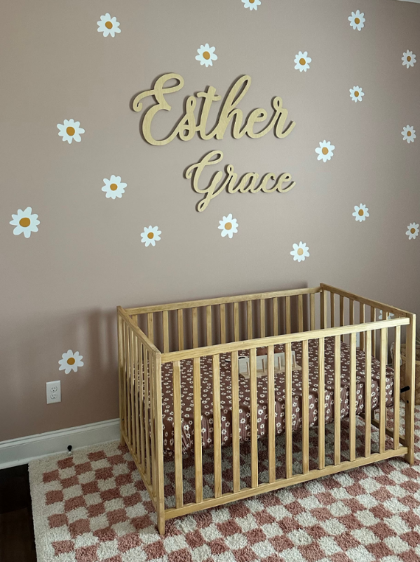 First and middle baby name wood sign over baby crib. The nursery is for a girl and there are daisies painted on the wall with a checkered rug. 