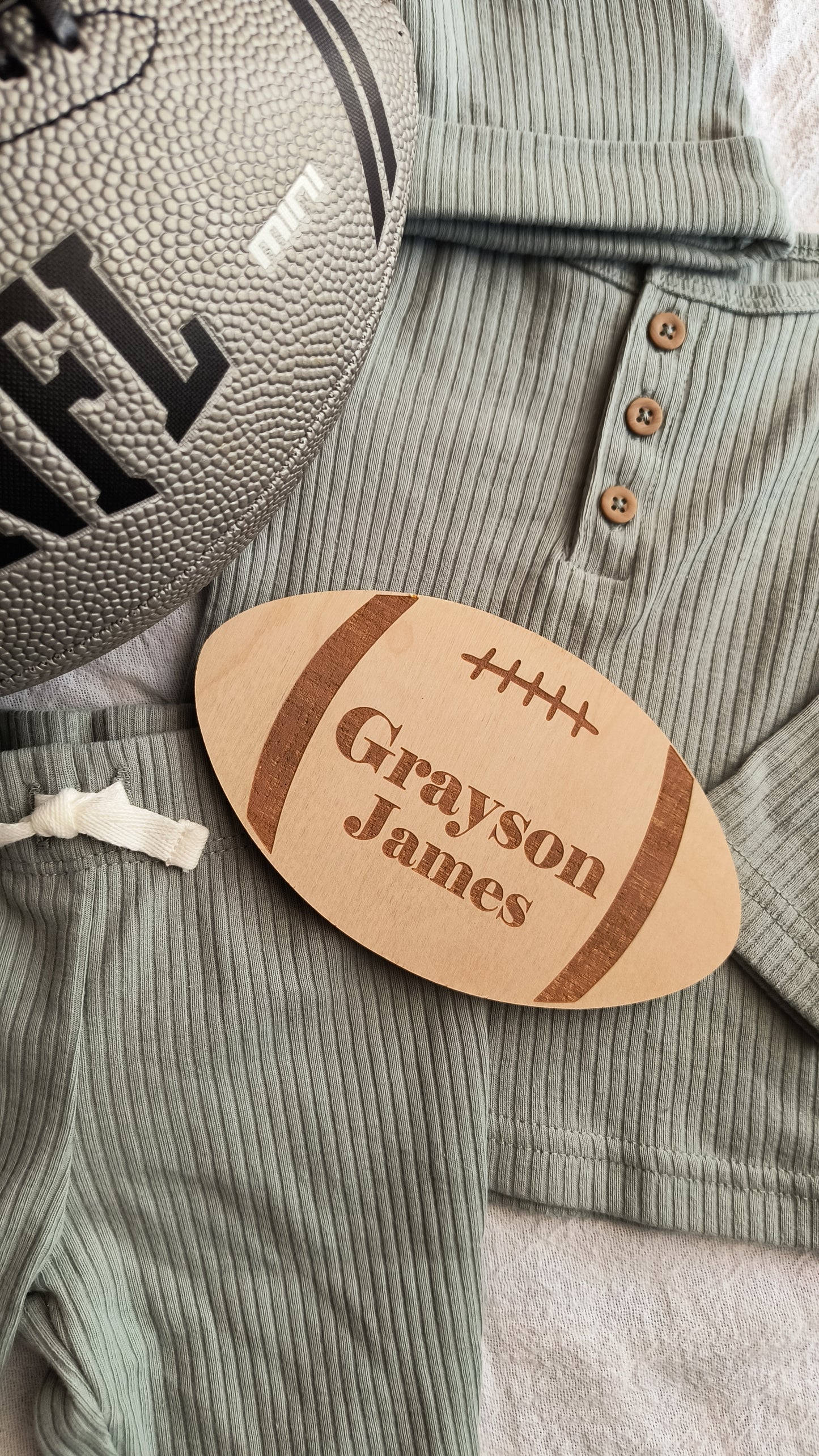 Football shaped engraved wood sign with football lacing and stripes engraved on the sign. It has a baby's first and middle name engraved as a baby announcement sign. There is a baby outfit and NFL football as props.