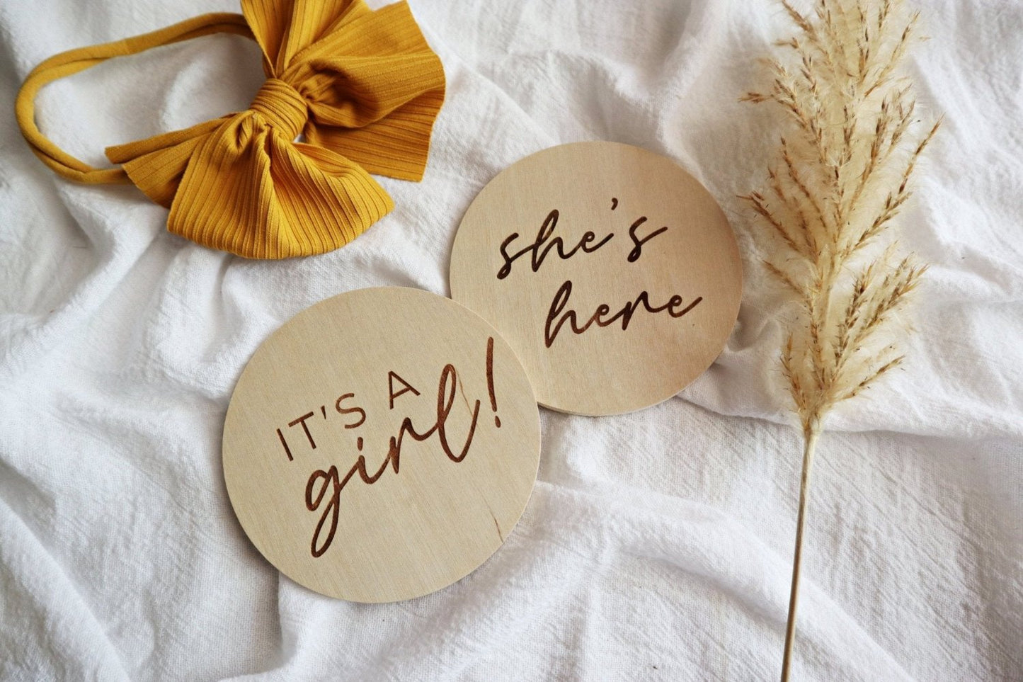 Flatlay of two engraved wooden signs on a white linen blanket with baby bow. One sign reads 'It's a girl!' and the other reads 'She's here.' Perfect for announcing a newborn’s arrival.