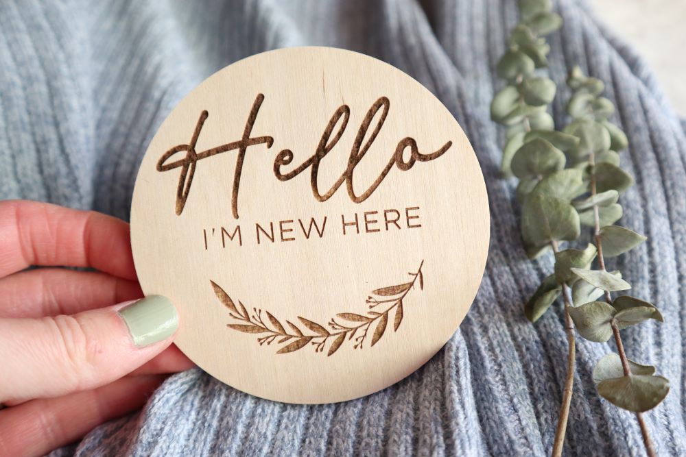 Hand holding engraved 'Hello I'm New Here' round wood sign. There is a blue baby blanket in the background and eucalyptus leaves to the right side. 