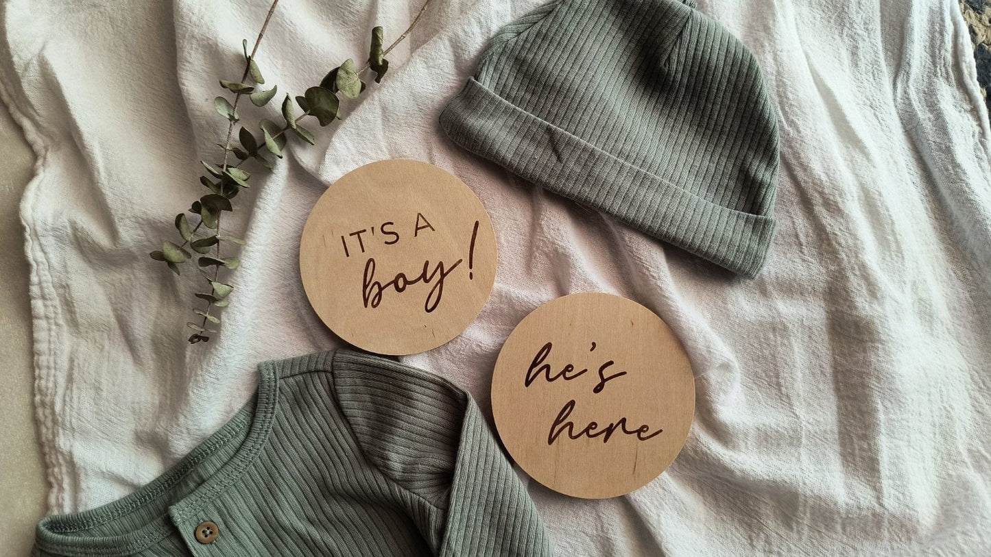 Two engraved wood signs with the phrases 'It's a Boy!' and 'He's Here!' laying on a soft white blanket, styled with an olive green baby outfit and eucalyptus leaves, perfect for baby boy announcements and newborn photo shoots.