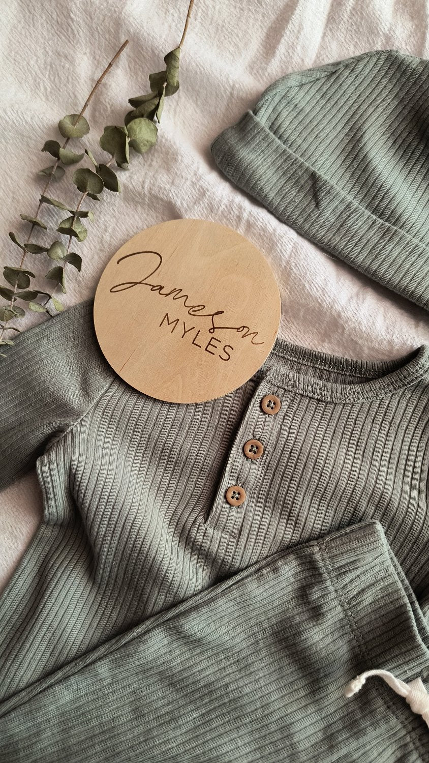 Engraved small wood baby name sign for a boy. The sign is on a flat lay with a sage green baby boy outfit and eucalyptus leaves.