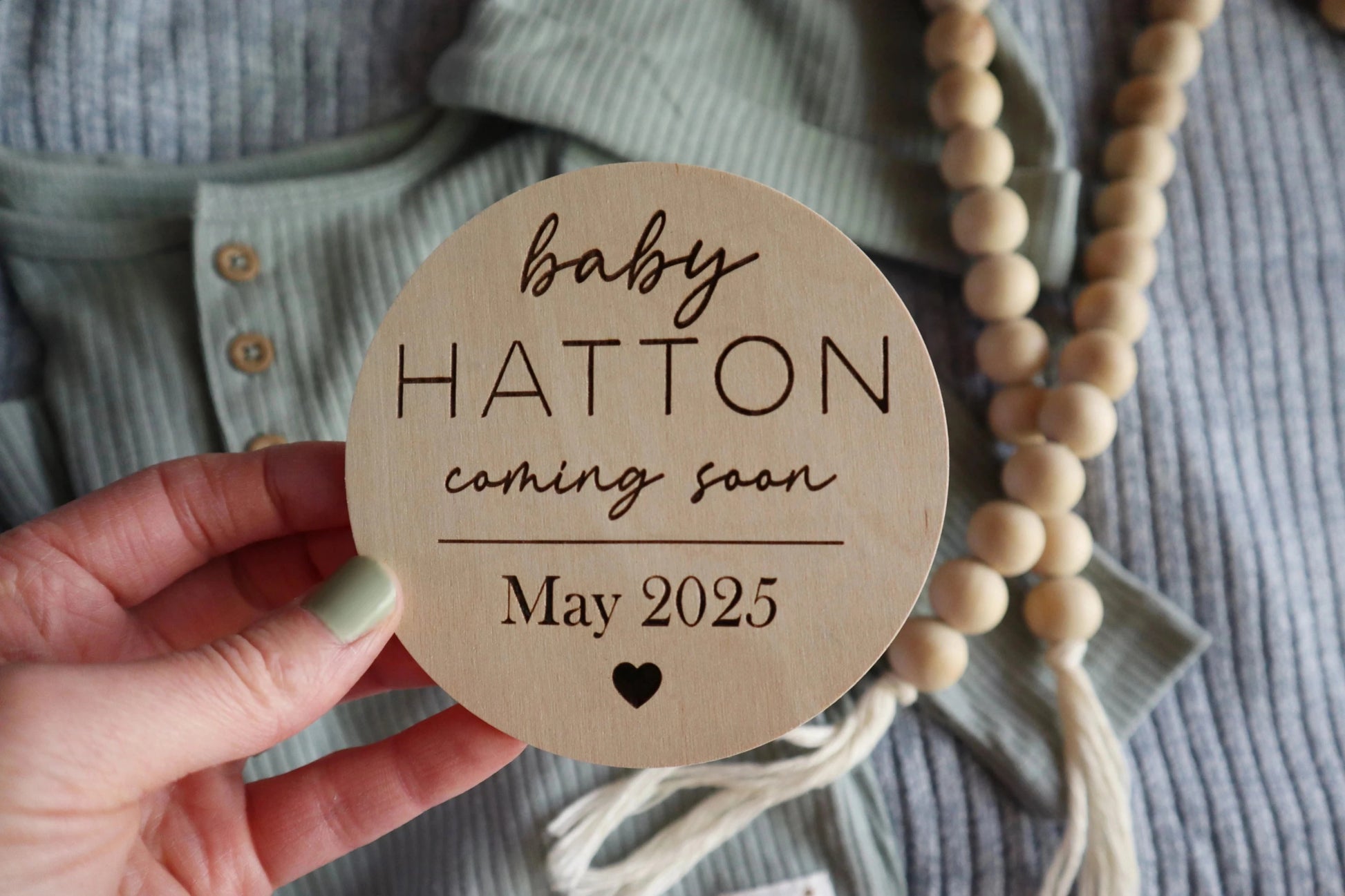 Hand holding round engraved baby announcement sign with baby outfit and wood beads in the background. The sign has baby's last name, coming soon, and the due date month and year. There is a heart engraved at the bottom of this sweet sign.